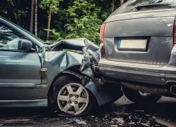 Two cars involved in a collision on a road surrounded by greenery. The front of a blue car is severely damaged, with the hood crumpled and the wheel bent. The rear of a gray SUV has collided with the front of the blue car, causing significant damage to both vehicles. Debris is scattered on the road beneath the cars