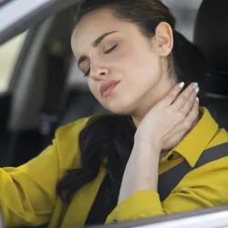 A person wearing a yellow shirt is seated in a car, with their hand on their neck, suggesting discomfort or pain. The seatbelt is fastened across their chest.